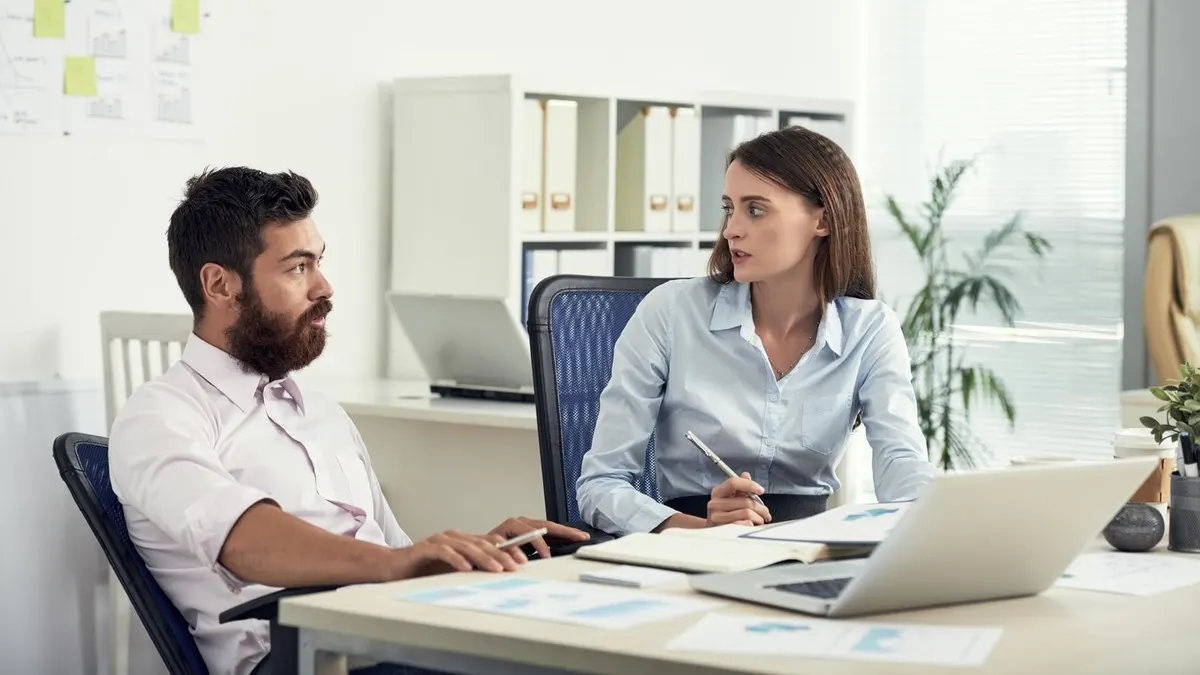 Two colleagues talk across a desk with a laptop and printed charts in a bright office