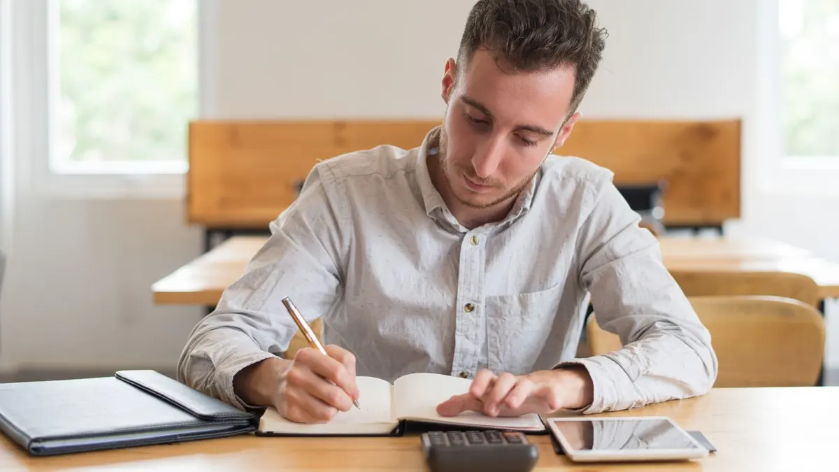 A young man writing notes in a notebook at a wooden desk in a bright study room
