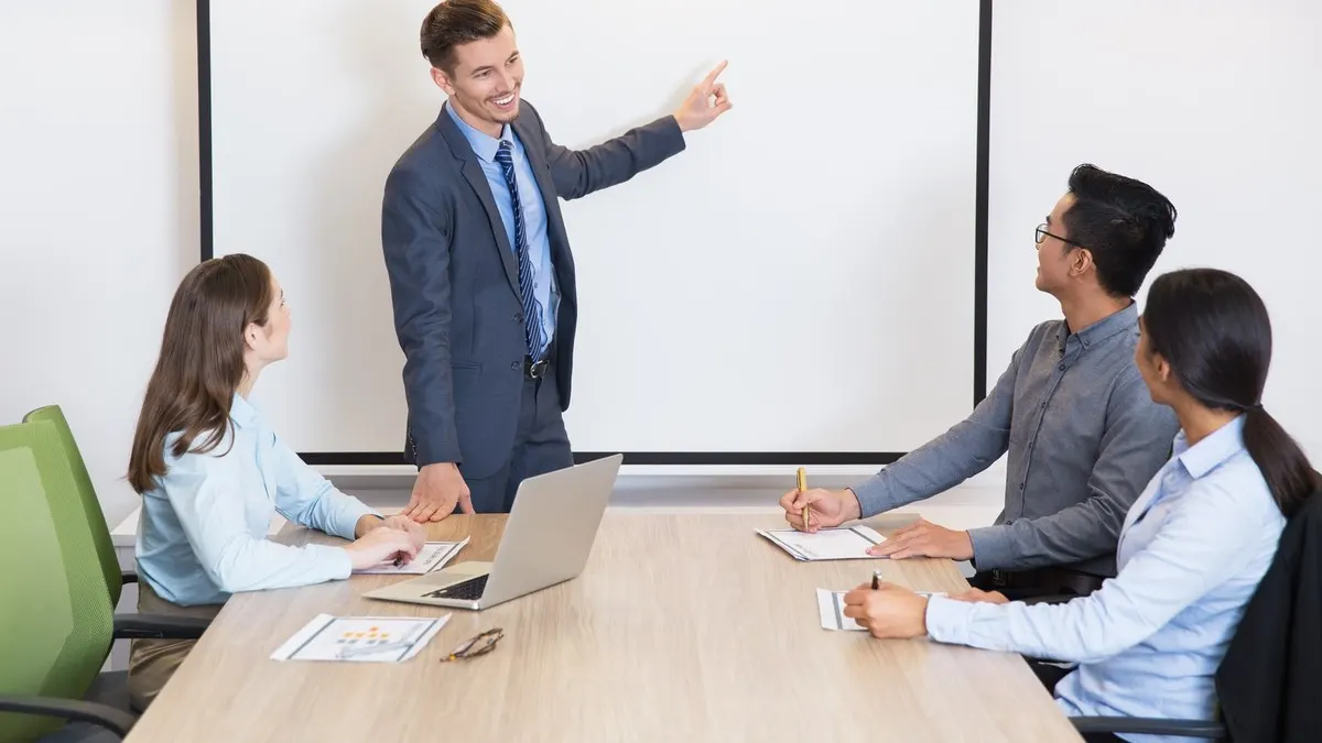 A man in a suit points to a blank screen while three colleagues at a meeting table listen