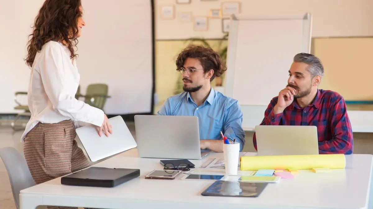 A woman joining two colleagues seated at laptops around a meeting table in a bright office