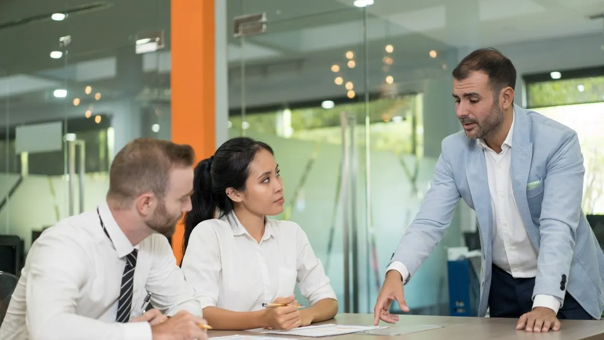 A manager in a light blue jacket leans over a meeting table and points at papers while two colleagues listen.