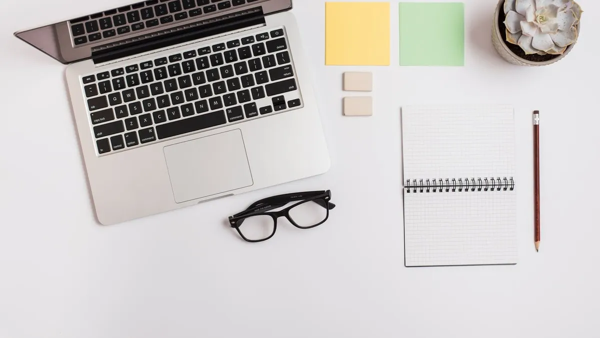 A laptop, a spiral notebook, sticky notes and glasses on a white desk
